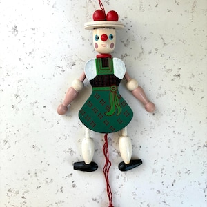 May include: A vintage wooden pull-string doll with a traditional folk design. The doll has a white hat with red accents, a green dress, and articulated limbs. The doll's face is painted, and it hangs from a red string.