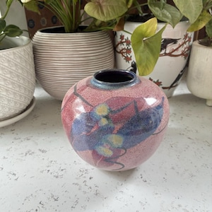 May include: A spherical ceramic vase with a speckled pink base glaze, featuring a prominent blue and yellow leaf-like pattern on the front. The vase has a smooth, glossy finish and a dark, narrow circular opening at the top with a deep blue rim.