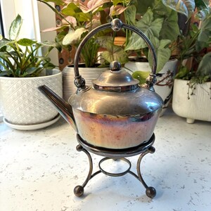 May include: A silver-toned metal teapot with a rounded body, a curved spout, and a high arched handle. The teapot rests on a matching three-legged metal stand with ball feet. The metal surface shows signs of tarnish and iridescent discolouration.