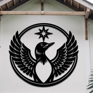 Könnte beinhalten: Ein schwarzes Metall-Wandkunstwerk mit einem stilisierten Vogel mit ausgebreiteten Flügeln, der in einem kreisförmigen Rahmen eingeschlossen ist. Über dem Vogel befindet sich ein Stern. Das Kunstwerk ist an einer weißen Wand montiert.