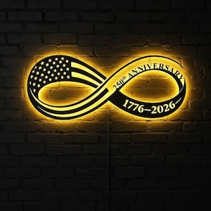 May include: A black metal sign in the shape of an infinity symbol, with the American flag on one side. The sign is illuminated with a warm yellow glow and features the text "250th Anniversary 1776-2026". The sign is mounted on a dark brick wall.