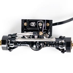 以下が含まれることがあります： 黒い金属製のRCクローラー用フロントアクスルアセンブリ。25Tサーボホーン付きサーボ、銀色のステアリングリンケージ、斑点模様のデフハウジングを備えています。黒いブラケットと固定用のネジが露出しています。