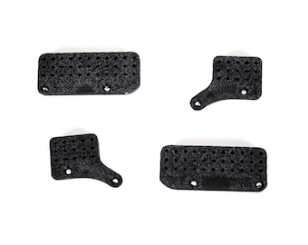May include: Four black, 3D-printed components with a textured, matte finish. Two pieces are rectangular with rounded corners and small circular holes, while the other two are L-shaped with similar perforated patterns and mounting points.