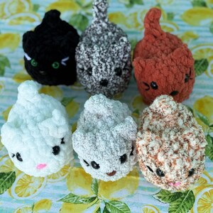 May include: Six handmade crochet cat toys in various colors: black, gray, orange, white, light gray, and brown. Each cat has embroidered facial features, including eyes and a nose. The toys are arranged on a lemon-themed fabric.