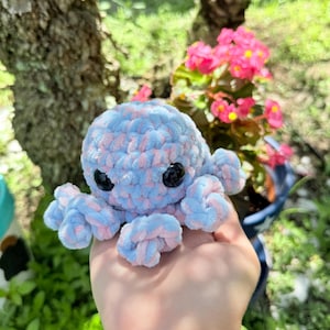 May include: A handmade crochet octopus in light blue and pink. The plush toy has black safety eyes and eight curled tentacles. The octopus is held in a hand, with a blurred background of greenery and pink flowers.