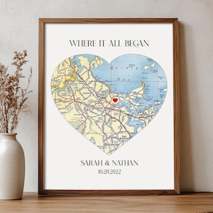 May include: A framed print with a heart-shaped map of a coastal area. The map is in shades of blue, green, and yellow, with a red heart marking a specific location. The text "WHERE IT ALL BEGAN" is at the top of the print, and "SARAH & NATHAN 10.28.2022" is at the bottom.