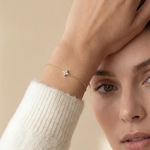 Puede incluir: Una delicada pulsera de oro con un pequeño diseño floral de cuatro pétalos con detalles brillantes. La pulsera se lleva en la muñeca, mostrando sus finos detalles y su elegante sencillez. La joya es un accesorio sutil pero llamativo.