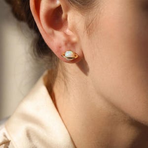 May include: A close-up of a person's ear wearing a gold-colored stud earring. The earring features a white opal stone with a gold ring around it, resembling a planet. The background is a soft, neutral color.