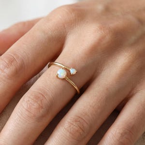 May include: A gold-colored ring with two opal gemstones. The ring has a delicate, swirling design, with one larger opal and a smaller one. The ring is on a person's finger.
