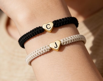 Könnte beinhalten: Zwei passende Armbänder mit goldenen Herzanhängern, jeweils mit einem einzelnen Buchstaben. Ein Armband ist schwarz, das andere beige. Die Armbänder sind aus geflochtenem Kordel und werden am Handgelenk getragen.