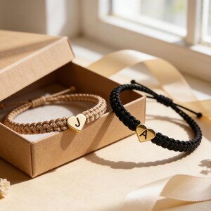 May include: Two macrame bracelets with gold heart charms, one in tan with a "J" and one in black with an "A", are shown. The bracelets are presented with a gift box and ribbon, suggesting a personalised gift.
