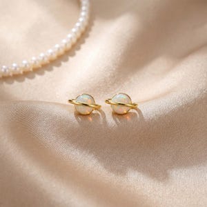 以下が含まれることがあります： 金色のスタッドピアス2つ。それぞれ白いオパール球が細い金の帯で囲まれています。ピアスはサテンの背景に置かれ、左上にはパールのネックレスが一部見えています。