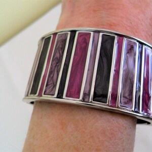 May include: A wide, silver cuff bracelet featuring vertical panels in purple, black, and grey. The bracelet, worn on a wrist, has a glossy finish. The design creates a striking contrast of colours and textures.