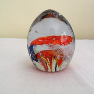 May include: Clear glass paperweight in an egg shape. Inside, there's a colourful design of red, white, yellow, and blue abstract forms. The paperweight is displayed on a white surface.