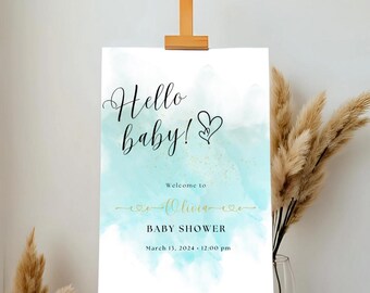 Pode incluir: Uma placa de boas-vindas para chá de bebê em um cavalete. A placa apresenta as palavras "Hello baby!" em escrita preta, com um fundo em aquarela em tons de verde-azulado e dourado. A placa também inclui o nome "Olivia" e a data e hora do chá de bebê.
