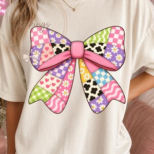 Puede incluir: Una camiseta color hueso con un gráfico central grande de un lazo hecho de un mosaico de patrones coloridos, que incluyen corazones rosas, margaritas blancas sobre fondo morado, cuadros vichy verdes, estampado de vaca y cuadros rosas y blancos.