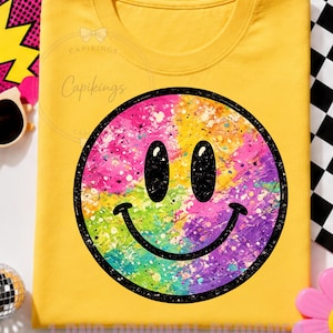 Puede incluir: Una camiseta de color amarillo brillante con un gráfico circular grande de una cara sonriente en el centro. La cara sonriente tiene un contorno negro, ojos ovalados negros y una boca curva, rellena con un diseño de salpicaduras multicolor.