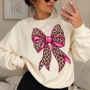 Pode incluir: Uma camisola de gola redonda cor de creme com um grande gráfico central de um laço. O design do laço combina uma cor rosa choque vibrante com um padrão de leopardo castanho e preto, exibindo fitas longas e fluidas que caem.