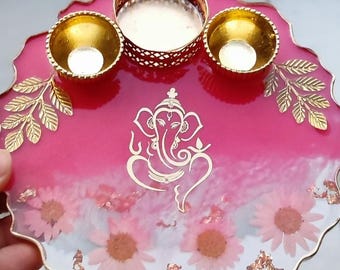 May include: A decorative pink resin tray with gold accents, featuring a gold Ganesh design and three gold candle holders. The tray is adorned with dried flowers and gold leaf details, perfect for home decor.