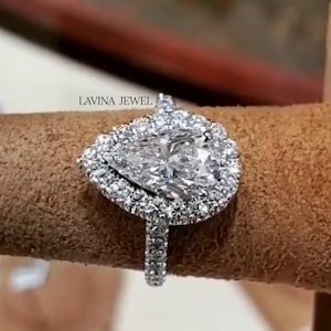 May include: A silver-toned ring featuring a large pear-shaped center stone surrounded by a halo of smaller round stones. The band is also encrusted with small round stones. The text LAVINA JEWEL is visible above the ring setting.