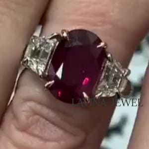 Puede incluir: Un anillo de plata con un gran rubí de talla ovalada como pieza central. El rubí es de color rojo intenso. Dos gemas rectangulares más pequeñas flanquean el rubí. El anillo está en un dedo con una banda dorada y esmalte de uñas rojo.