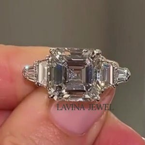 Op de afbeelding: Een platina ring met een grote, vierkant geslepen centrale diamant, geflankeerd door twee kleinere, rechthoekige diamanten. De ring heeft de tekst "LAVINA JEWEL" op de band. De ring wordt door een persoon vastgehouden.