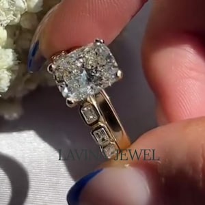 May include: A gold ring featuring a large, rectangular-cut diamond. The ring is held up by a hand with blue nail polish. The band has smaller stones. The background includes white flowers and the text "LAVINA JEWEL".