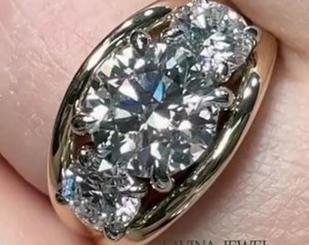 Può includere: Un anello d'oro con tre grandi diamanti scintillanti. L'anello ha una fascia arrotondata e un'incastonatura che tiene saldamente i diamanti. I diamanti sono chiari e riflettono la luce, creando una brillantezza. Il testo "LAVINA JEWEL" è visibile.