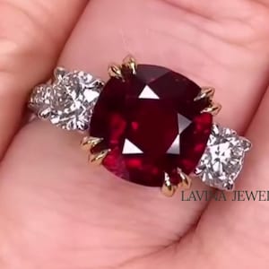 Könnte beinhalten: Nahaufnahme eines Rings mit einem großen, quadratisch geschliffenen, tiefroten Edelstein, flankiert von zwei kleineren, klaren Edelsteinen. Der Ring hat eine goldene Fassung um den roten Stein und ist mit "LAVINA JEWEL" markiert.