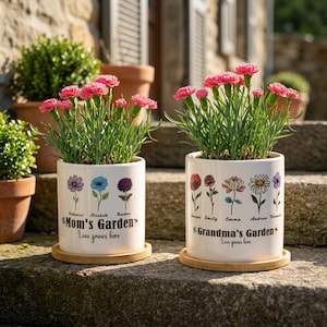 May include: Two white ceramic flower pots with pink carnations. Each pot has a floral design and the words "Mom's Garden" and "Grandma's Garden" with the phrase "Love grows here". The pots sit on bamboo saucers.