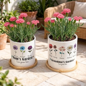 May include: Two white ceramic flower pots with pink carnations. One pot reads "Mom's Garden" and the other reads "Grandma's Garden." Each pot has a wooden saucer. The pots are decorated with floral illustrations and names.