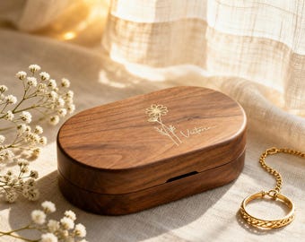 May include: An oval-shaped dark brown wooden box with a smooth finish. The lid features a gold-colored engraved line drawing of a daisy-like flower and the name Victoria in cursive script. The box is closed and has a simple, clean design.
