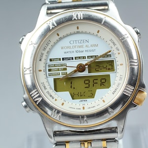 Könnte beinhalten: Silber-goldene Citizen Worldtime Alarm-Uhr mit weißem Zifferblatt. Die Uhr zeigt Uhrzeit, Datum und Alarmeinstellungen an. Sie hat eine Digitalanzeige und analoge Zeiger. Wasserdicht bis 10 bar.