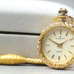 Puede incluir: Un reloj de bolsillo dorado con esfera marfil y agujas doradas. El reloj tiene la palabra "QUARTZ" impresa en la esfera y la marca "TENSHODO". Está sujeto a un cordón trenzado dorado.