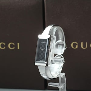 Könnte beinhalten: Rechteckige Gucci-Uhr mit silberfarbenem Gehäuse und schwarzem Zifferblatt mit sich wiederholendem Muster. Die Uhr hat ein weißes Lederarmband und wird auf einem transparenten Ständer präsentiert. Der Hintergrund zeigt das Gucci-Logo.
