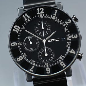 May include: A black Seiko chronograph watch with a round face, featuring large white Arabic numerals from 1 to 12, three sub-dials, a tachymeter scale, and a date window at the 3 o'clock position. The watch has a black metal band and silver hands.
