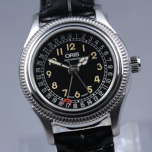 Peut inclure: Une montre automatique Oris argentée avec un cadran noir et des chiffres dorés. La montre dispose d'un affichage de la date et d'un bracelet en cuir noir. Le cadran affiche le nom de la marque et le mot "AUTOMATIC".