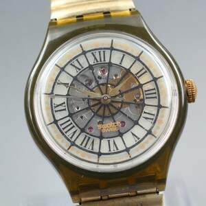 May include: A close-up of a Swatch watch with a transparent olive green case and a gold-tone metal band. The watch face features Roman numerals, a skeleton design, and the word "SWISS". The watch hands are gold-tone.