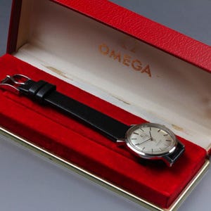 May include: A vintage Omega wristwatch with a silver dial and black leather strap, presented in a red box. The watch features silver hour markers and hands. The box is red with gold lettering.