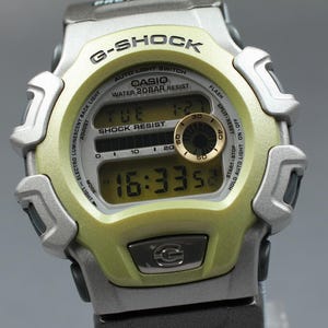 May include: A G-Shock digital watch with a light green and silver case. The watch face displays the time, date, and other functions. The watch has a black strap and is water-resistant.
