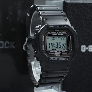 May include: Black G-Shock digital watch with a rectangular face displaying the time. The watch has a black band and casing. The watch face shows the time as 14:35:22. The watch is labeled with the text "G-SHOCK".