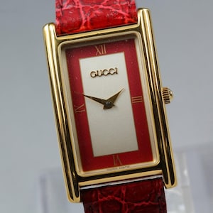 Puede incluir: Reloj Gucci rectangular dorado con esfera roja y blanca y correa de cuero roja. La esfera del reloj presenta el logotipo de Gucci y agujas doradas. La correa es roja texturizada. Números romanos marcan las horas.