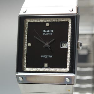 Puede incluir: Un reloj Rado Diastar Quartz plateado con una esfera cuadrada negra. La esfera del reloj tiene un borde con incrustaciones de diamantes y muestra la fecha. El reloj tiene una correa plateada y está marcado "Swiss Made".