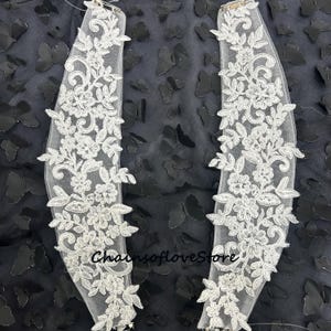 Puede incluir: Dos apliques de encaje blanco con diseños florales y de hojas. Los apliques están sobre un respaldo de malla transparente. El fondo es una tela oscura con detalles de mariposas. El texto "ChainsofloveStore" es visible.