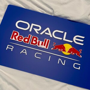 以下が含まれることがあります： 青い長方形のサインで、Oracle Red Bull Racingのロゴが特徴です。ロゴには、白と赤の文字で「ORACLE」、「Red Bull」、「RACING」という文字が含まれています。2匹の赤い雄牛が黄色の円の上に描かれています。