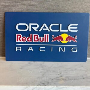 以下が含まれることがあります： 青い長方形の看板。白い文字で「ORACLE」、2頭の赤い雄牛と黄色い太陽が描かれた赤と白のレッドブルのロゴ、そして白い文字で「RACING」と記されている。看板は暗い背景に対して立体的に浮き上がったデザインとなっている。