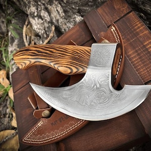 May include: A handcrafted axe with a curved blade and wooden handle. The blade features intricate engraved designs and is paired with a brown leather sheath. The axe rests on a wooden box, showcasing its craftsmanship.