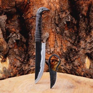 May include: A handcrafted black knife with a bird-shaped handle and a leather-wrapped grip. The blade is dark with a silver edge. A smaller, matching knife is also present. The knives are displayed on a wooden surface against a textured brown background.