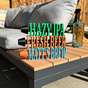 May include: A wooden beer flight with the words "HAZY IPA", "FRESH BEER", and "MATT'S BREW" in green, orange, and blue. The flight has three black holders for glasses and sits on a wooden table. A bottle is in the background.