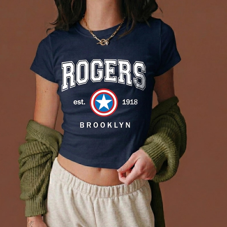 Peut inclure: T-shirt bleu marine avec le mot "ROGERS" en lettres blanches. En dessous, un embl&egrave;me circulaire avec une &eacute;toile bleue et l'inscription "est. 1918 BROOKLYN". Le t-shirt est &agrave; manches courtes.